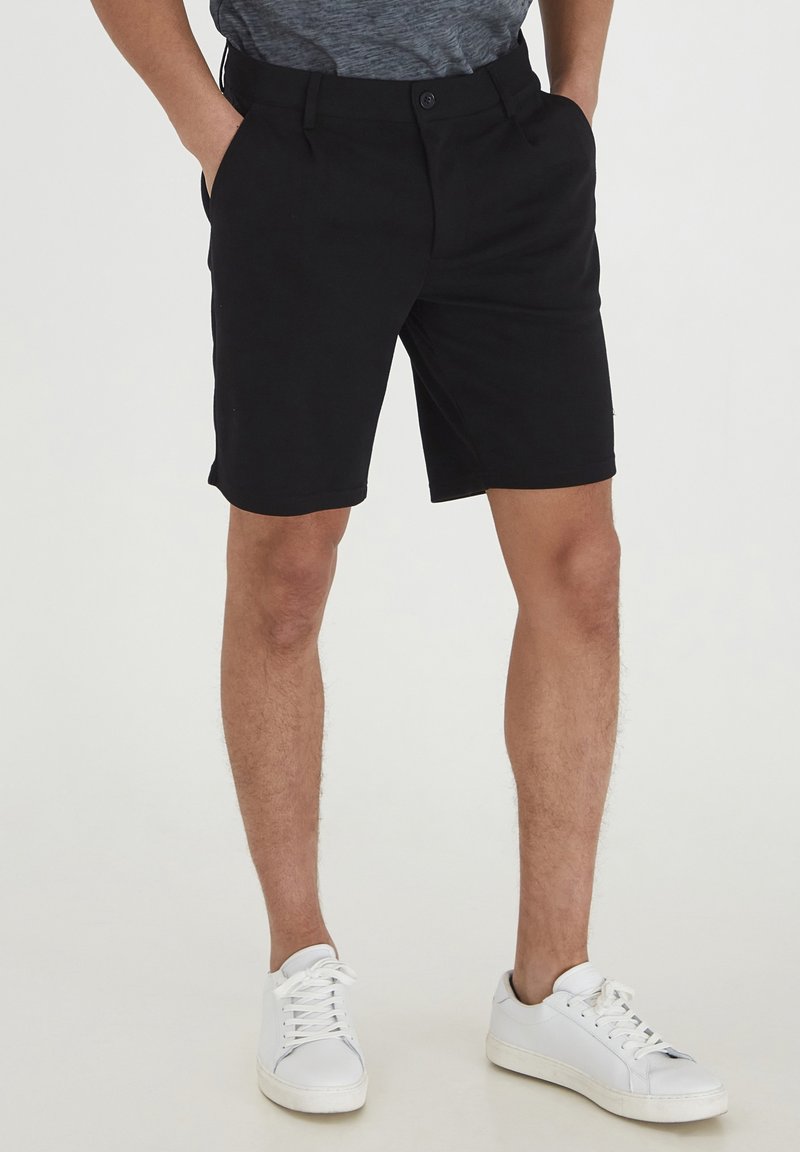 Homme portant un short noir sur mesure, une chemise grise rentrée, et des baskets blanches basses, debout les mains dans les poches.