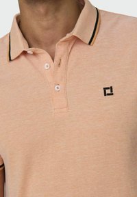 Polo de tejido ligero en melocotón claro con un patrón texturizado; presenta un collar a rayas negro y naranja, tres botones y un pequeño logo negro.