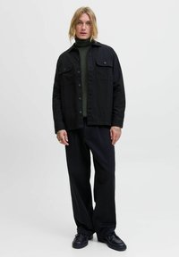 Schwarze Jacke mit zwei Brusttaschen, darüber ein dunkelgrüner Rollkragenpullover. Weite schwarze Hose und schwarze Schuhe komplettieren das Outfit.