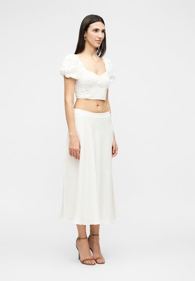 Femme debout portant un top court blanc à manches bouffantes avec broderie florale et une jupe midi blanche taille haute ainsi que des talons beiges à bride à la cheville.