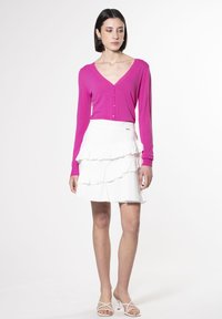 Cardigan fucsia a maniche lunghe e gonna bianca a strati con volant. Dettagli a volant sulla gonna, texture liscia e silhouette aderente. Sandali bianchi con cinturini.