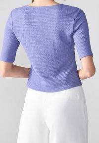 Světlý fialový texturovaný top s kulatým výstřihem a třičtvrtinovými rukávy, kombinovaný s bílými, přiléhavými kalhotami.