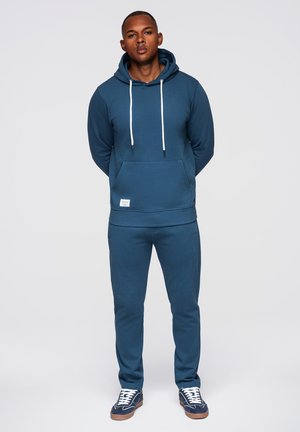 Mann steht mit den Händen hinter dem Rücken, trägt einen blauen Hoodie, passende Jogginghose und blaue Sneaker vor einfachem Hintergrund.