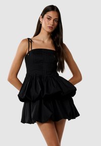 Robe noire avec un corsage sans bretelles ajusté et une jupe évasée à volants. Présente une texture plissée et des bretelles ajustables. Tissu lisse.