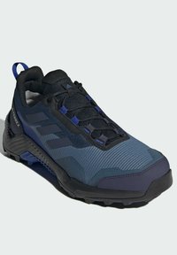 Blå och svart vandringsko med en texturerad ovandel, förstärkt tåhuvud, gummisula och platta snören. Har tre mörka ränder på sidan.
