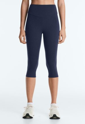 Kobieta w granatowych krótkich legginsach i białych tenisówkach, stojąca na tle jednolitego białego tła.