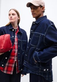 Γυναίκα με κόκκινο πουκάμισο καρό και σκούρο τζιν μπουφάν κρατώντας κόκκινη τσάντα, άνδρας με σκούρο τζιν μπουφάν και μπεζ καπέλο με το χέρι στην τσέπη.