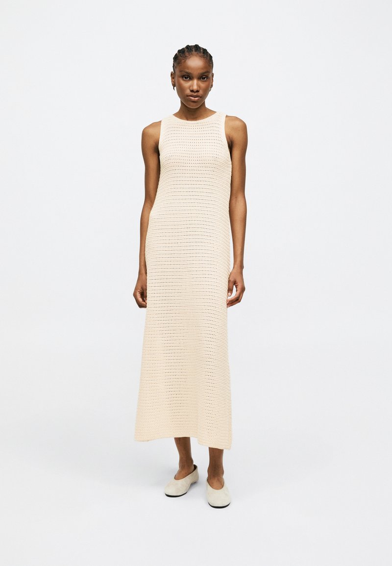 Femme debout portant une robe tricotée beige sans manches, longueur jusqu'aux chevilles, et des chaussures légères à enfiler, sur fond blanc uni.