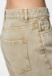 Ingrandimento di pantaloni in denim beige che mostrano la tasca posteriore, i passanti della cintura e la cucitura laterale sulla vita e fianchi di una persona.