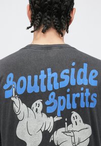 Pelēka T-krekls ar lielu zilu tekstu "Southside Spirits" un divu baltu spoku ilustrāciju, kas tur sveces.