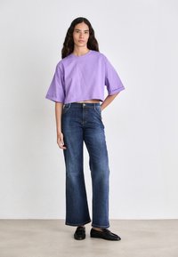Purple cropped t-shirt with short sleeves paired with dark blue wide-leg jeans. Model wearing black slip-on shoes, standing against a neutral background.