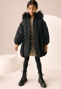 Black puffer jacket with a faux fur hood, elastic cuffs, and cinched waist. Worn over a leopard print dress and black leggings, with lace-up boots.
