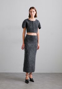 Vrouw in een fluffy donkergrijze korte mouwen cardigan met knopen, bijpassende lange rok, zwarte puntige hakken en een parelketting.
