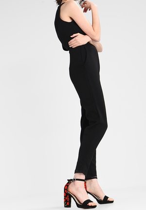 Kobieta w czarnym, bezrękawowym kombinezonie i czarnych sandałach na wysokim obcasie z czerwonym, kwiatowym haftem na masywnych obcasach, stojąca bokiem.