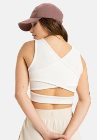 White cropped top with crisscross design at the back, made of textured fabric. Paired with beige, loose-fitting, light pants and a brown cap.