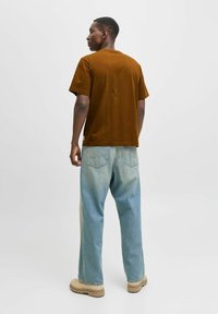 Camiseta de algodón marrón con mangas cortas, combinada con unos jeans de mezclilla azul claro. Los zapatos son beige. El fondo es completamente blanco.