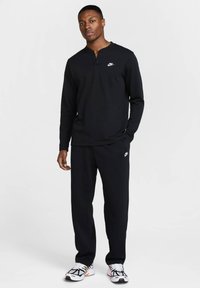 Melnas garām piedurknēm henley krekliņš ar Nike logo, apvienots ar melniem sporta biksēm, kurām ir balts Nike logo, valkāts ar baltām un melnām sporta apaviem.