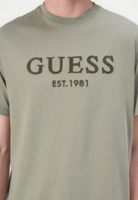 Maslinasto zeleni pamučni majica s kratkim rukavima, s uzdignutim vezenim tekstom "GUESS" i "EST. 1981" u tamnije zelenoj boji. Glatka tekstura.