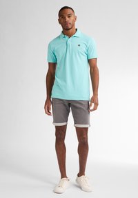 Polo azzurro con colletto, maniche corte e un piccolo logo sul petto, abbinato a pantaloni corti grigi con orlo arrotolato e scarpe da ginnastica bianche.
