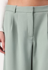 Pantalons vert clair en tissu texturé avec une taille haute, des plis à l'avant et des poches latérales. Design ajusté avec une finition élégante.