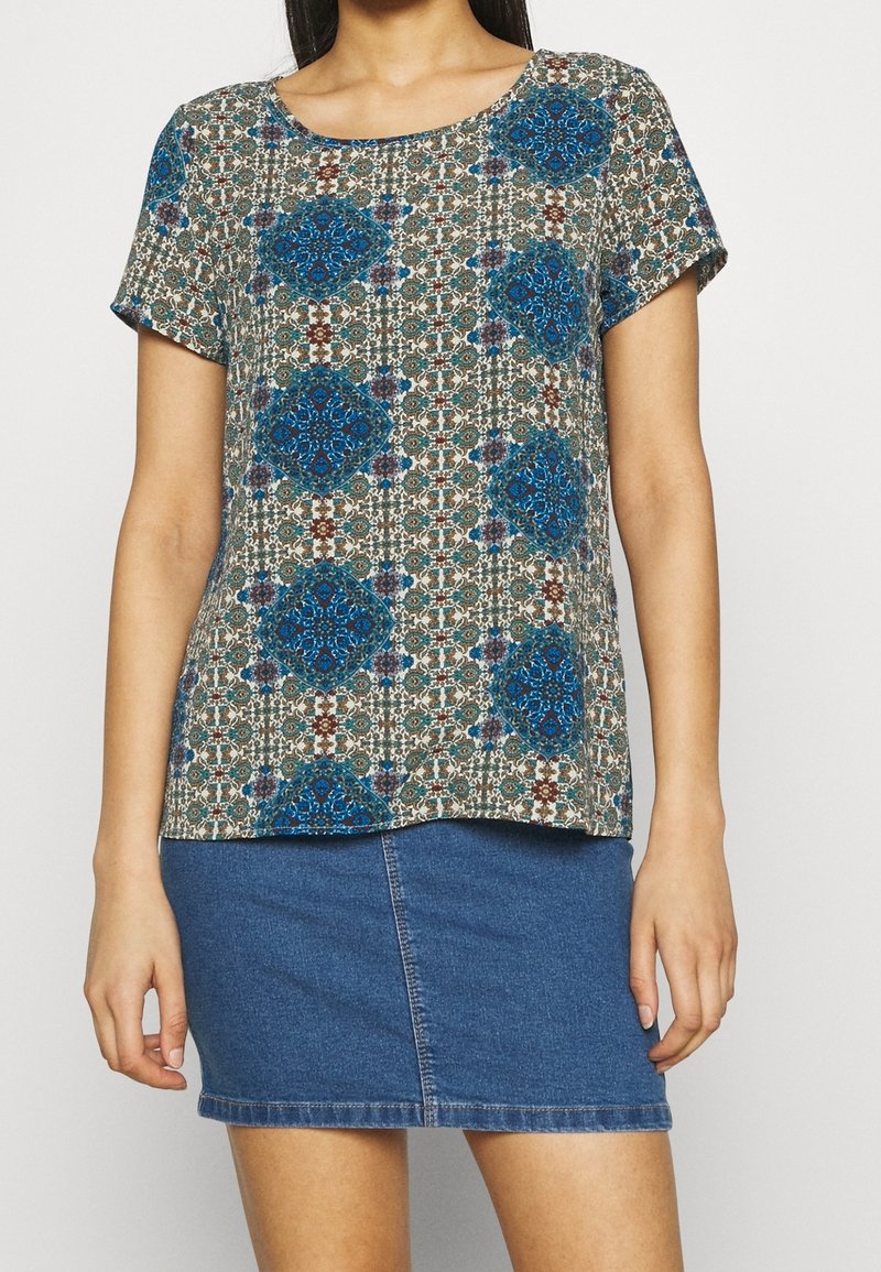 Short-sleeve top with a patterned design in blue, brown, and cream on a light background; round neckline and loose fit. Paired with a denim skirt.