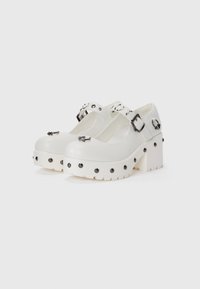 White platform shoes with buckle straps, rounded toes, and a textured sole. Accents include black studs around the base and silver hardware.