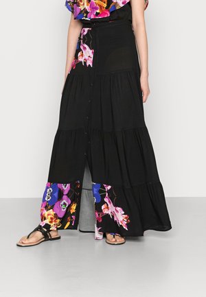 Femme portant une longue jupe noire avec des motifs floraux violets et roses près de l'ourlet et au milieu, assortie à des sandales noires.