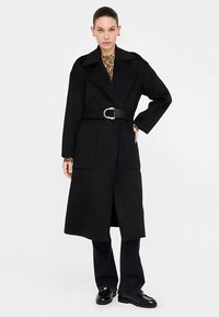 Schwarzer Mantel mit Gürtel, weitem Kragen, zwei Fronttaschen und glatter Textur. Kombiniert mit schwarzer Hose und Lederschuhen.