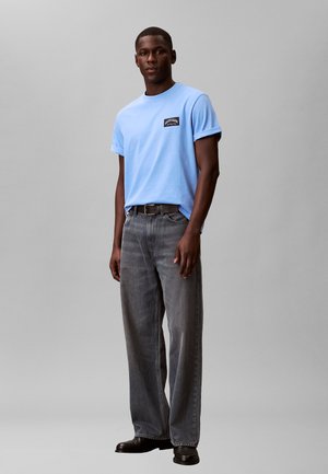 Mann, der ein hellblaues Kurzarmhemd, dunkelgraue Jeans, schwarzen Gürtel und schwarze Schuhe trägt, vor einem schlichten grauen Hintergrund steht.