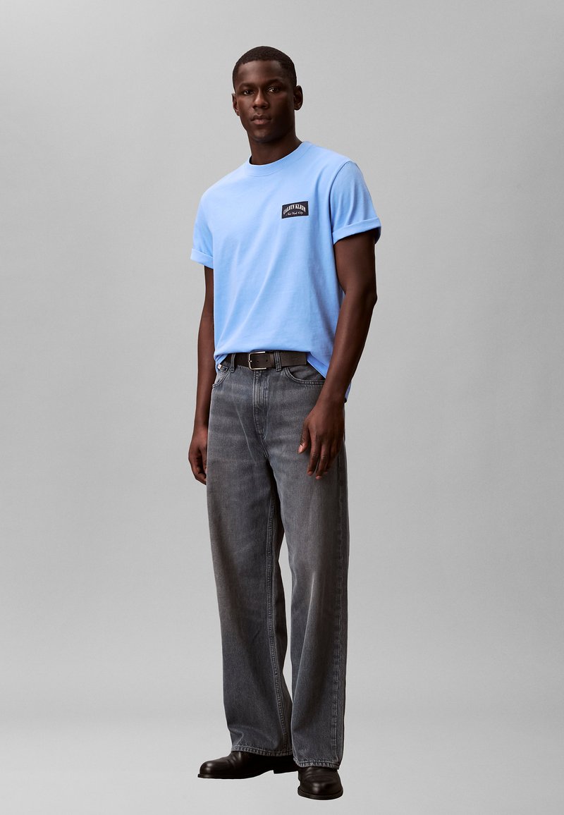 Homme portant une chemise à manches courtes bleu clair, un jean gris foncé, une ceinture noire et des chaussures noires, debout devant un fond gris uni.