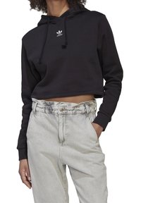 Svart kort hoodie med dragsko och en liten vit logotyp på bröstet, kombinerad med ljusgrå höga jeans med hög midja.