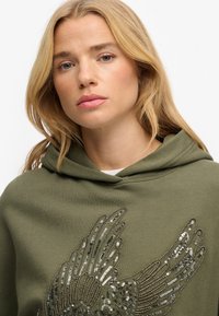 Felpa con cappuccio verde oliva con un grande motivo a ali di paillettes argento e dorate, dotata di cappuccio con coulisse e tessuto morbido e strutturato.