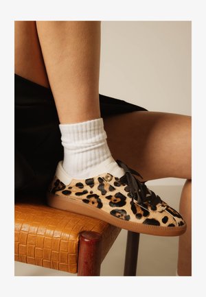 Jambes portant des chaussettes blanches et des baskets imprimé léopard reposant sur une chaise en cuir tressé orange avec pieds en bois.