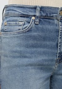 Κοντινό πλάνο ατόμου που φοράει ανοιχτόχρωμο τζιν με τσέπη για κέρματα, θηλιά για ζώνη και εμφανή μεταλλικά πριτσίνια· μπεζ ριμπ μπλούζα μεσα στη μέση.