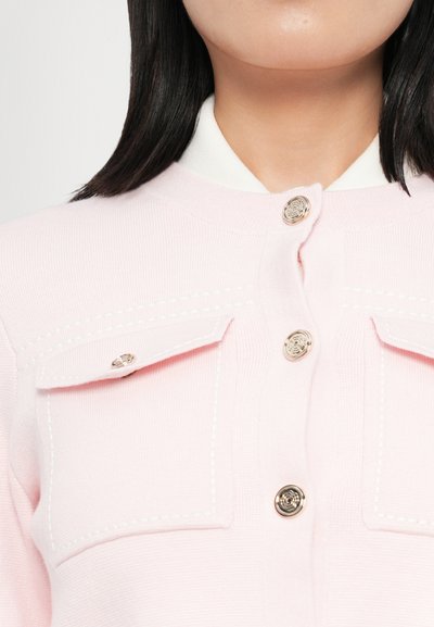 Světle růžový cardigan s kulatým límcem, stříbrnými knoflíky, dvojitými náprsními kapsami a bílými švy na kapsách.