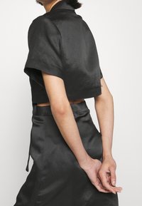 Svart satin kort topp med korta ärmar och hög krage. Designen har en slät textur och en minimalistisk stil.