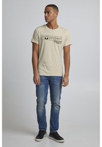 Beige bomull t-skjorte med svart grafisk tekst, rund hals, kort erm, kombinert med blå jeans og svarte joggesko.