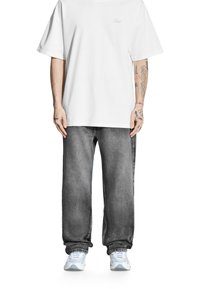 Biała, obszerna koszulka, szare luźne dżinsy i szare sneakersy. Znaczące tatuaże na ramionach. Koszulka ma delikatne logo; dżinsy mają teksturę denimu.