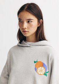 Sweatshirt à capuche gris clair avec un graphique d'orange et de citron en orange, jaune et violet, accompagné de feuilles vertes. Texture du tissu douce.