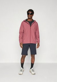 Waterbestendig roze jack met capuchon, gecombineerd met zwarte korte broek. Het model draagt een zonnebril en lichte sneakers, staand tegen een neutrale achtergrond.