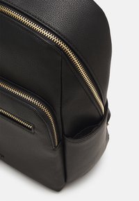 Mochila de piel negra con cremalleras doradas, múltiples compartimentos y un acabado texturizado, con bordes redondeados y un diseño elegante.