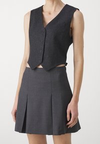 Dunkelgraue, taillierte Weste mit V-Ausschnitt, fünf Knöpfen und einer kleinen Tasche. Kombiniert mit einem grauen Faltenrock. Beide Teile sind maßgeschneidert.