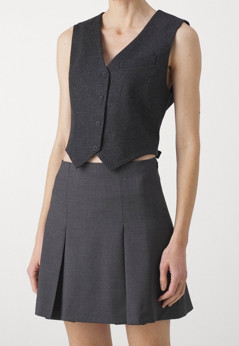 Dunkelgraue, taillierte Weste mit V-Ausschnitt, fünf Knöpfen und einer kleinen Tasche. Kombiniert mit einem grauen Faltenrock. Beide Teile sind maßgeschneidert.