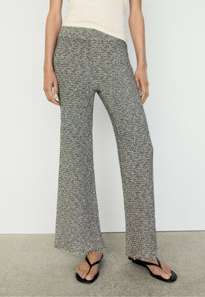Donna che indossa pantaloni in maglia grigi a vita alta e gamba larga con orli grezzi e sandali neri infradito, in piedi contro una parete chiara e semplice.