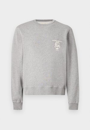 Mann mit lockigem Haar, der einen grauen Sweatshirt, hellblaue Jeans und schwarze Lederschuhe trägt, steht mit einer Hand in der Tasche.