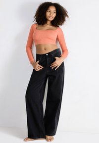 Młoda kobieta z kręconymi włosami, ubrana w koralowy długi rękaw crop top i szerokie czarne dżinsy, stoi boso na tle białym.
