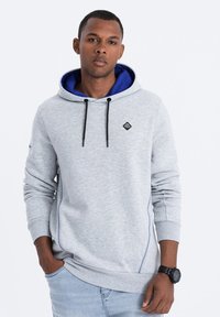 Sudadera gris con forro interior azul, cordones ajustables, acentos negros y un logo. Presenta costuras laterales y puños de canalé.
