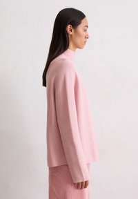 Lichtroze, geribbelde trui met hoge hals en lange mouwen, gecombineerd met een bijpassende geribbelde rok, gepresenteerd tegen een effen achtergrond.