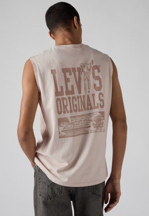 Homme portant un t-shirt beige sans manches Levi's Originals avec un grand logo et un graphisme au dos, associé à un jean gris foncé.