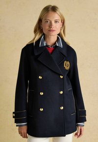 Tumes sinipesaga kulmusega kuldsete nööpidega ja tikandiga embleemiga. Omab topeltkinnitatud disaini, teravat kraed ja kontrastseid triibulisi varrukaotsi.